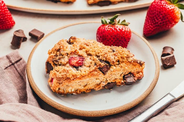 A chocolate strawberry scones on a white plate with chocolate chunks around it and fresh strawberries.