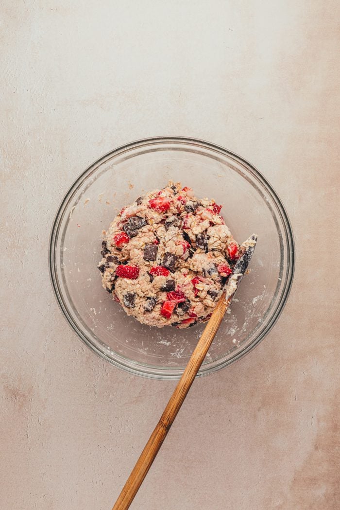 A glass bowl with the dough mixed with strawberries and chocolate and a spatula.