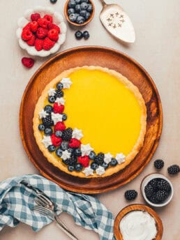 A classic lemon tart with berry and whipped cream garnishing it and bowls of berries in the background.