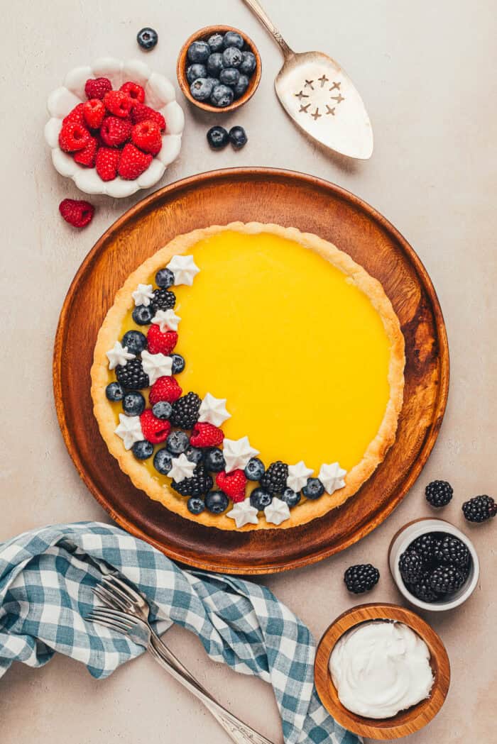 A classic lemon tart with berry and whipped cream garnishing it and bowls of berries in the background.