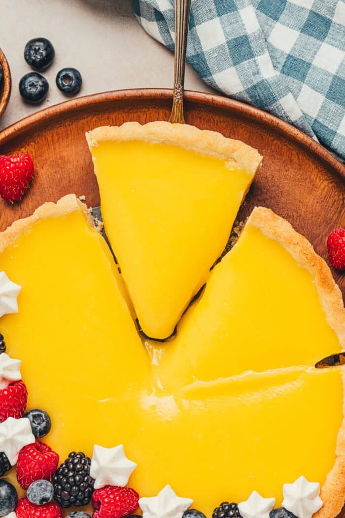 A homemade classic lemon tart on a tray, decorated with berries with a slice being removed.
