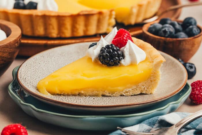 A slice of classic lemon tart on two plates, decorated with berries and whipped cream.