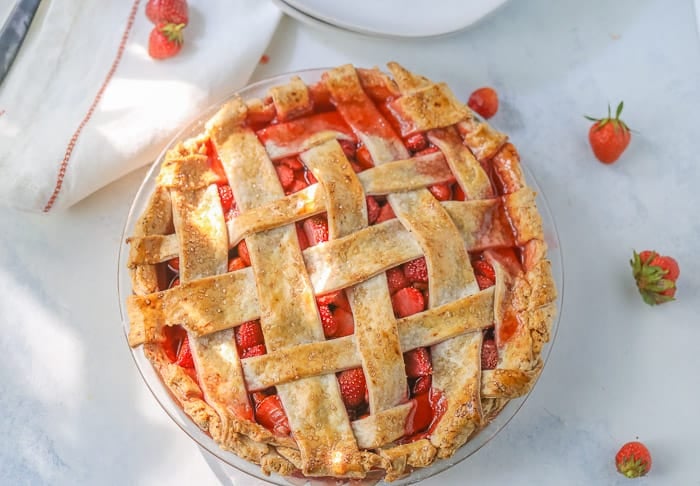 A picture of a homemade strawberry pie with a lattice pie crust and a few fresh strawberries on a table.
