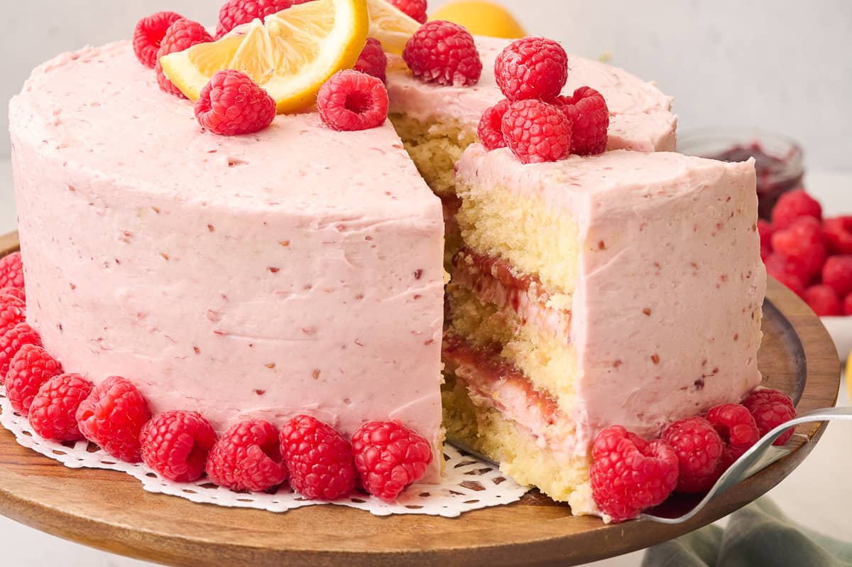 Lemon raspberry cake on a cake stand with a slice being removed.