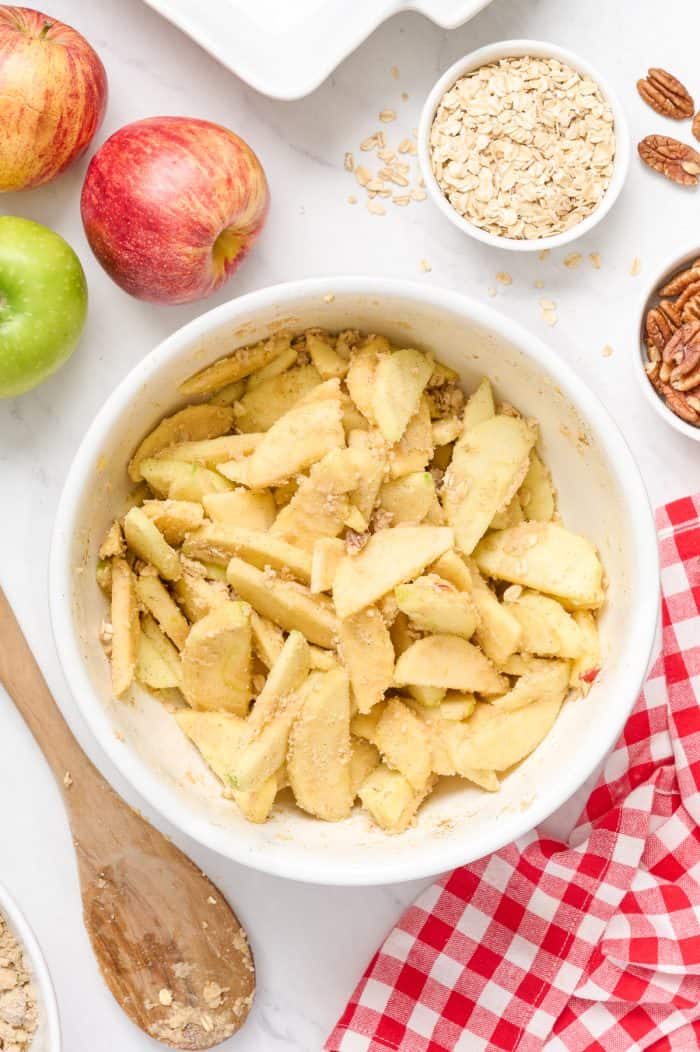 The sliced apples are tossed with some of the apple crisp topping in a white bowl.