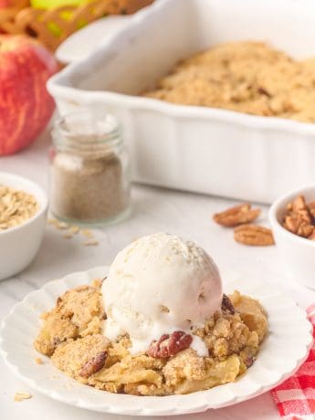 A plate filled with apple crisp with a scoop of ice cream and the tray of apple crisp in the background.