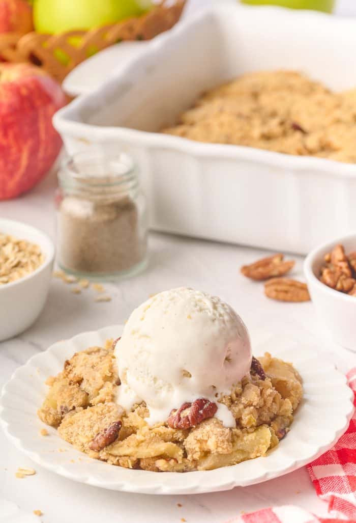 A plate filled with apple crisp with a scoop of ice cream and the tray of apple crisp in the background.