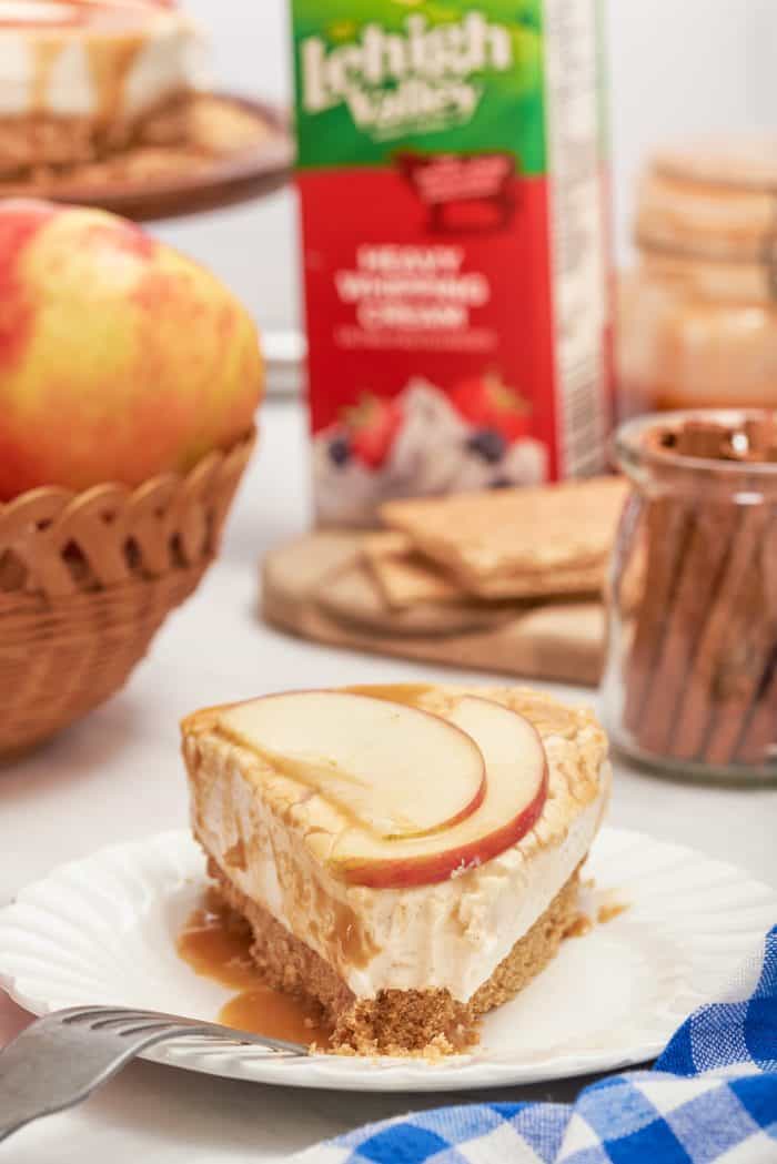 A closeup of a slice of apple cheesecake with a carton of heavy whipping cream in the background.