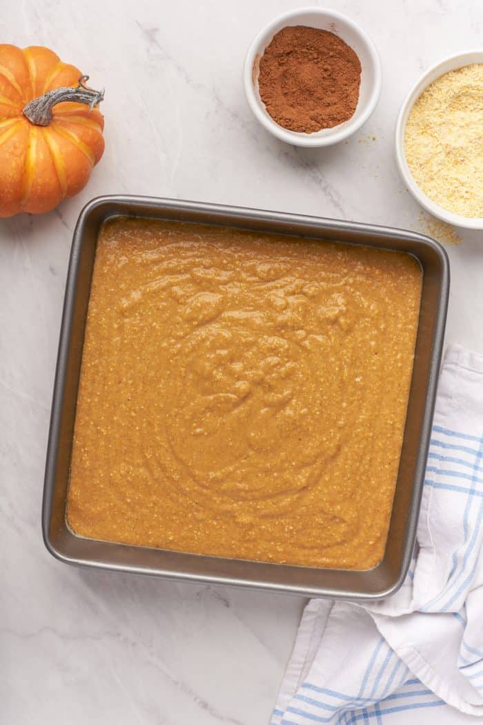 A baking pan filled with raw pumpkin cornbread batter with a small pumpkin and bowls of ingredients.