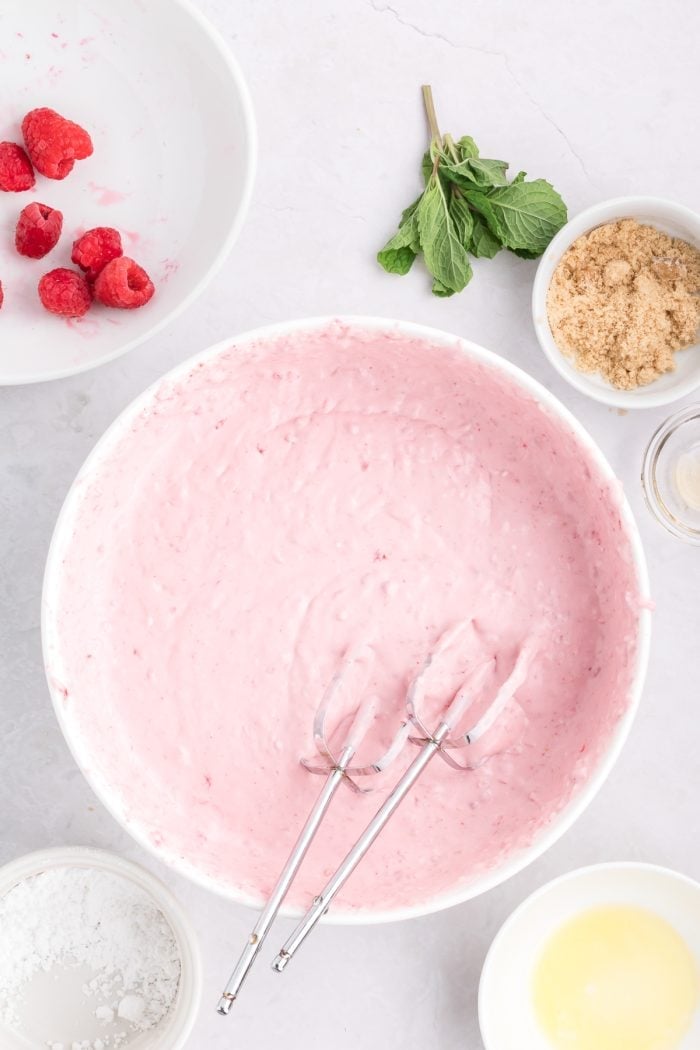 Whipping cream, cream cheese, and raspberries mixed together in a white bowl with a pair of beaters from a hand mixer. 