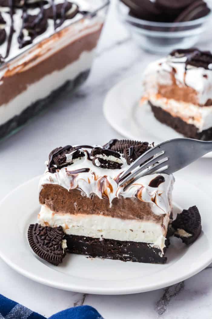 Two plates with slices of no bake Oreo layered dessert with the glass dish with the dessert in it in the background. One slice of dessert has a fork going into it.