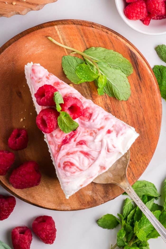 An overview of a slice of cheesecake with fresh raspberries and mint leaves on a wooden cutting board. 