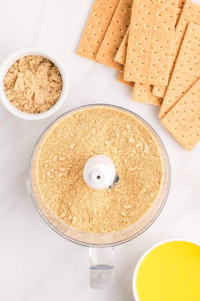 A food processor with graham cracker crumbs and graham crackers in the background.