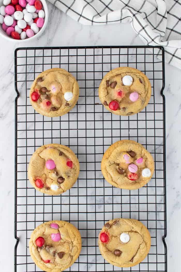 Six Valentine's M&M cookies cooling on a rack with a bowl of M&M in the background.