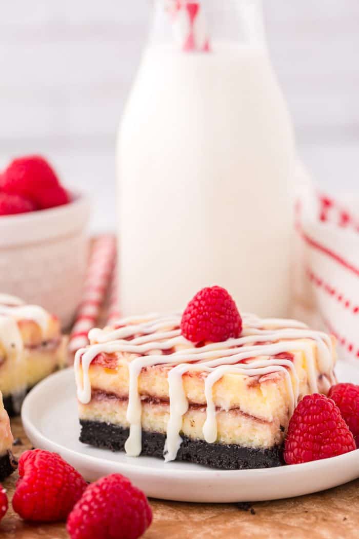 A white chocolate raspberry cheesecake bar on a white plate with a bottle of milk in the background and fresh raspberries. 