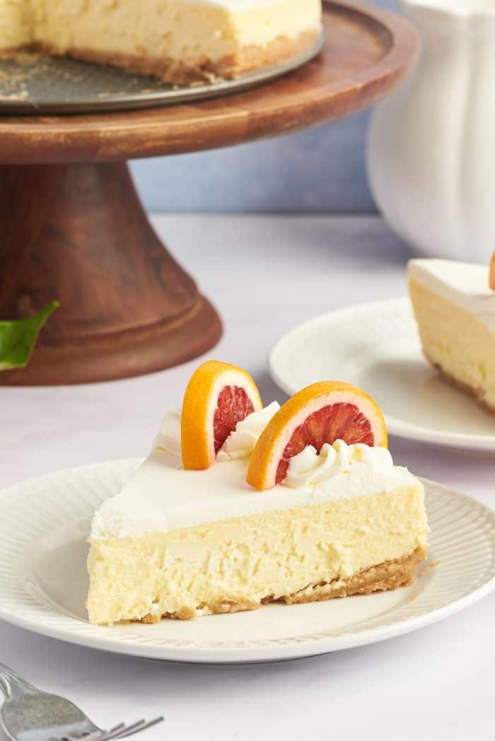 A slice of winter citrus cheesecake on a white plate with a wooden cake stand in the background. 
