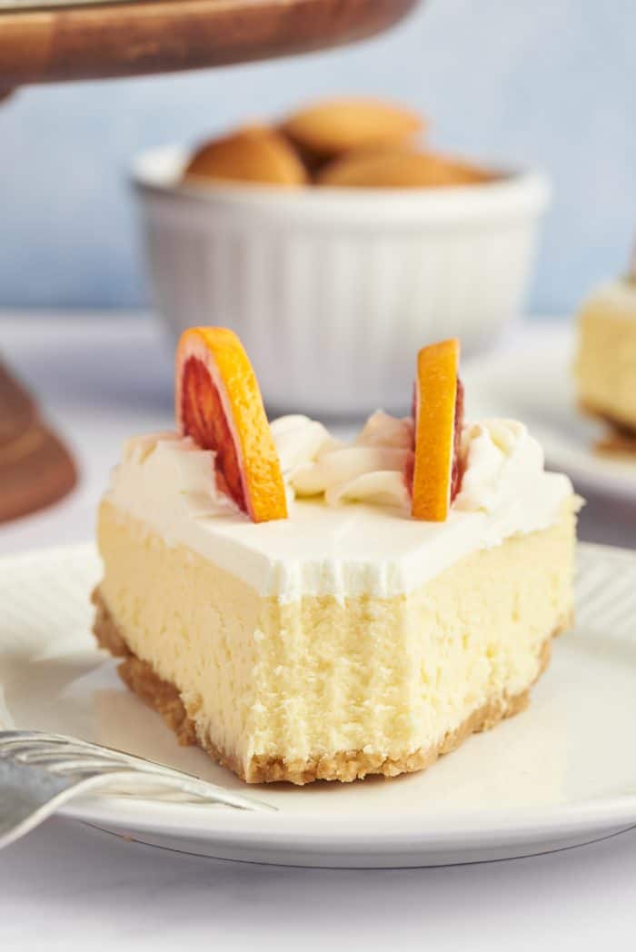 A slice of winter citrus cheesecake with a bite taken out of it on a white plate with a wooden cake stand in the background.