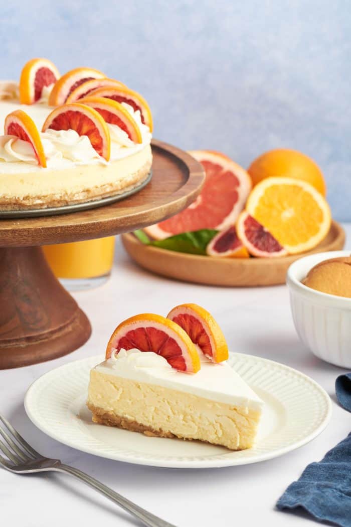 A slice of winter citrus cheesecake on a white plate with a wooden cake stand in the background.