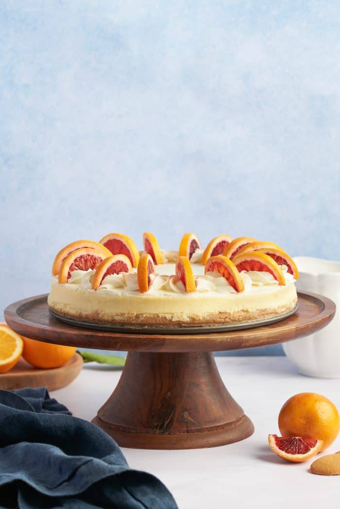 A blood orange cheesecake with sliced blood oranges on a wooden cake plate against a blue background. 