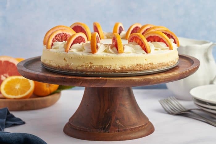 A blood orange cheesecake with sliced blood oranges on a wooden cake plate against a blue background.