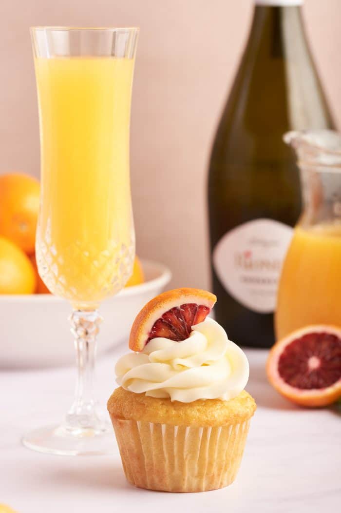 A mimosa cupcake with a glass of mimosa and a glass bottle of orange juice and Prosecco in the background.