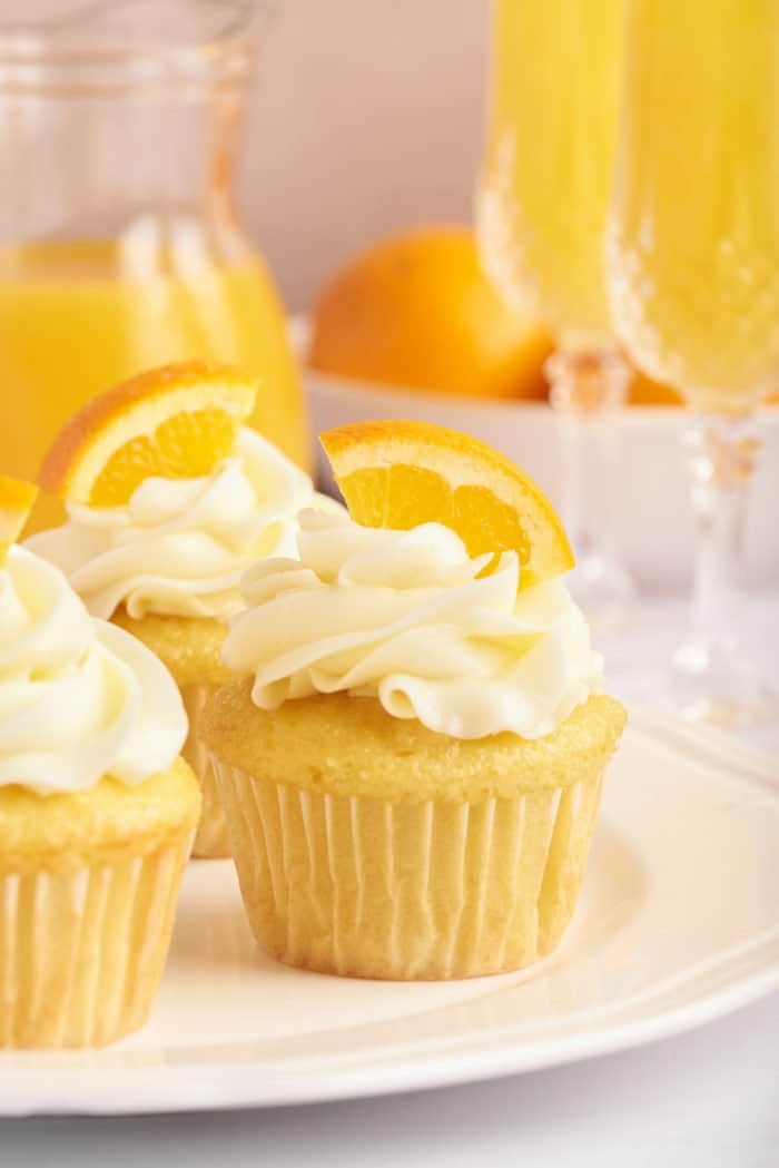 Mimosa cupcakes on a white plate with a glass of mimosa and a glass bottle of orange juice  in the background.