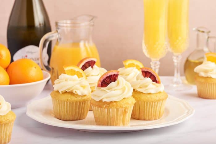 A white plate with various mimosa cupcakes on it and Prosecco, bottles of orange juice, and two glasses of mimosas. 