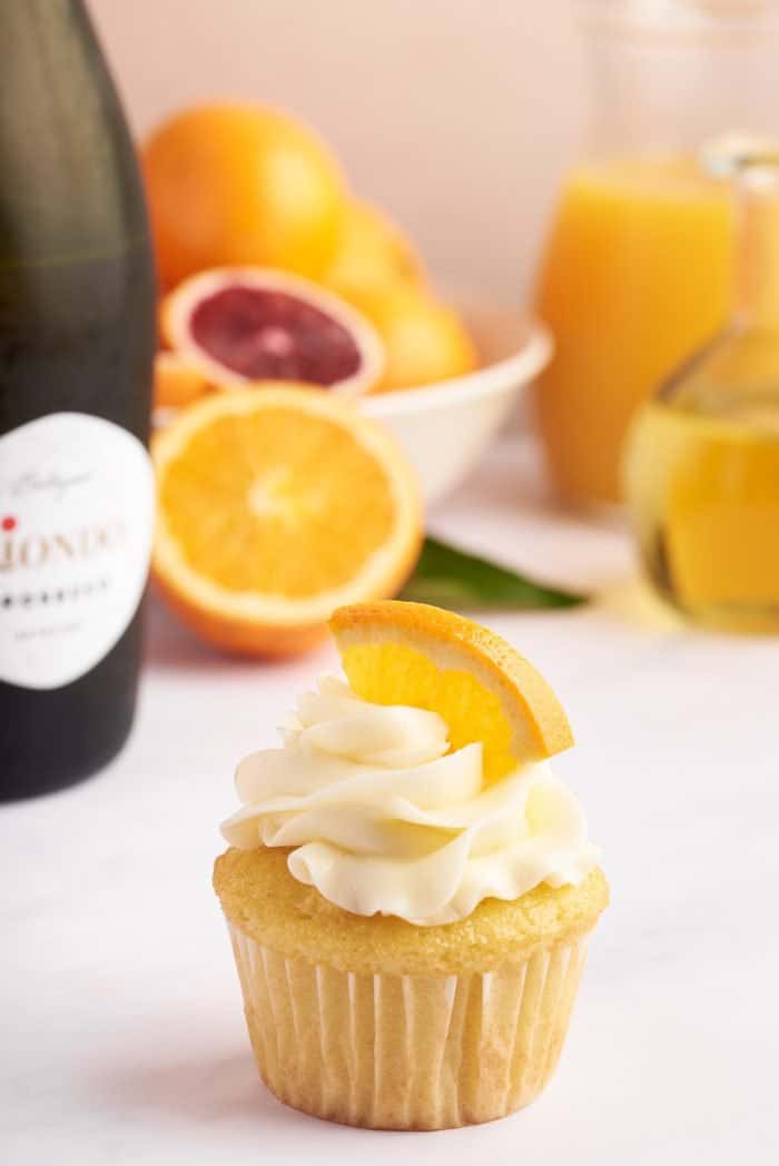 A closeup of a mimosa cupcake with a slice of orange and a bottle of Prosecco in the background.