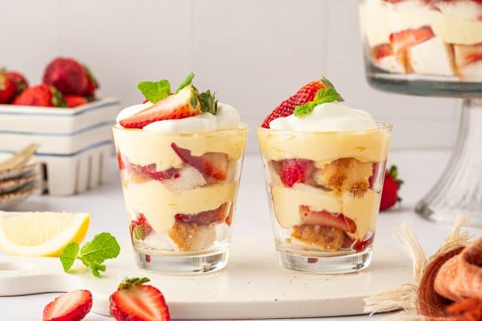 Two glass filled with strawberry trifle with strawberries and strawberry trifle in the background.