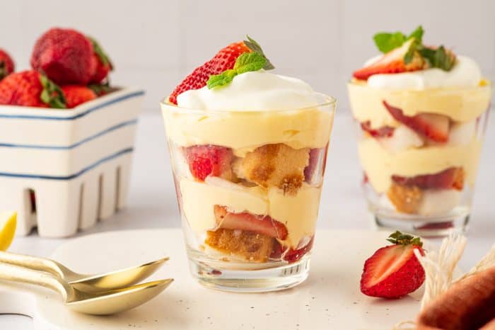 Two glass filled with strawberry trifle with strawberries in the background and gold spoons.