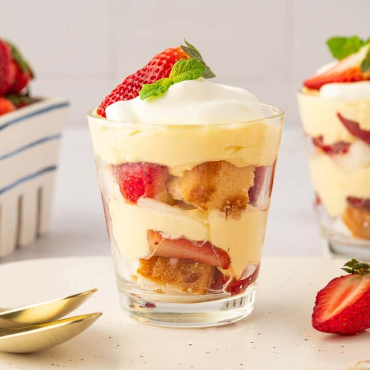 Two glass filled with strawberry trifle with strawberries in the background and gold spoons.