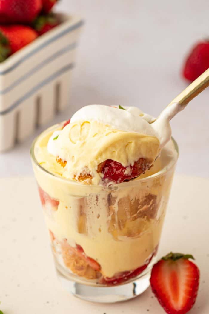 Two glass filled with strawberry trifle with strawberries in the background and a spoon scooping out a bite.