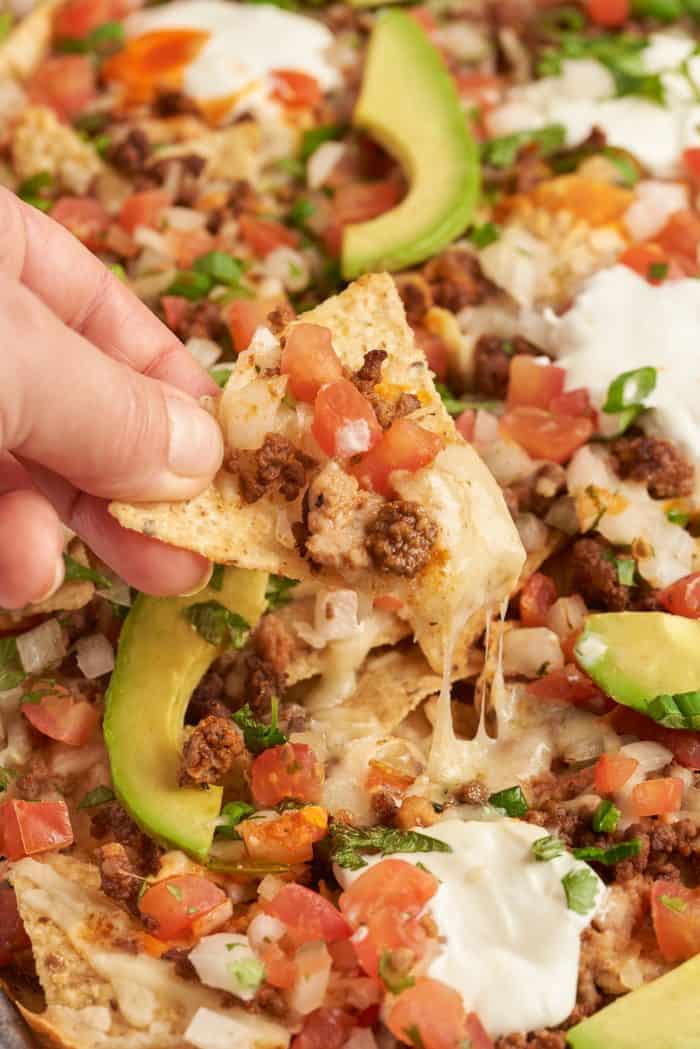 Baked sheet pan nachos with pico de gallo and sliced avocado. A hand is picking up a tortilla chip with melted cheese and toppings.