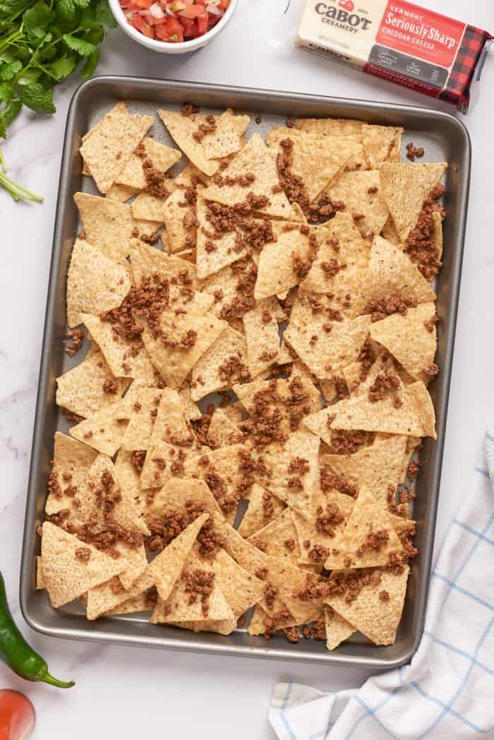 Ground beef added to tortilla chips on a baking pan.