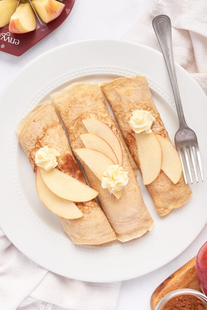 A white plate with 3 rolled crepes filled with cooked apples, and garnished with apple slices.