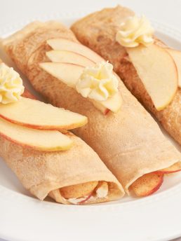 A white plate with three apple crepes that are filled with a cooked apple filling and garnished with sliced apples.