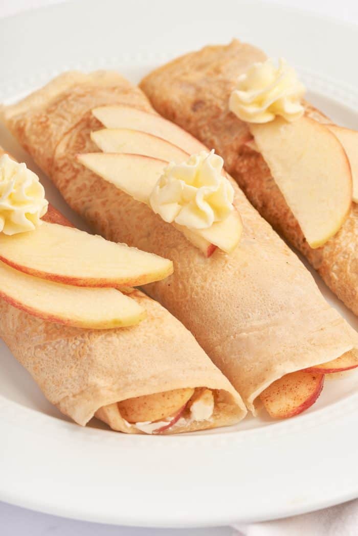 A white plate with three apple crepes that are filled with a cooked apple filling and garnished with sliced apples.