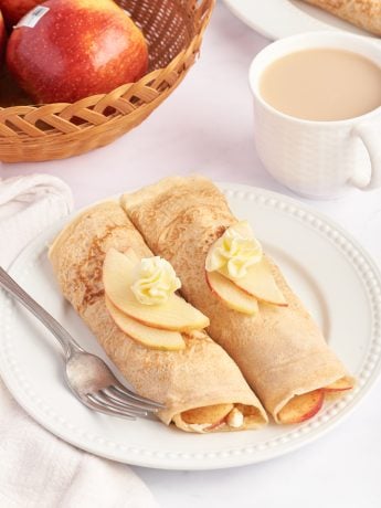 A white plate with two apple crepes that are filled with a cooked apple filling and garnished with sliced apples.
