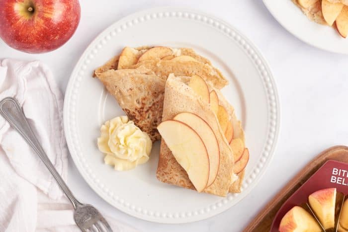 A white plate with two apple crepes that are filled with a cooked apple filling and garnished with sliced apples.