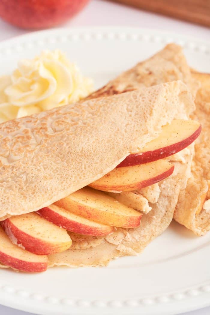 A white plate with a crepe filled with cooked apples, and garnished with apple slices.