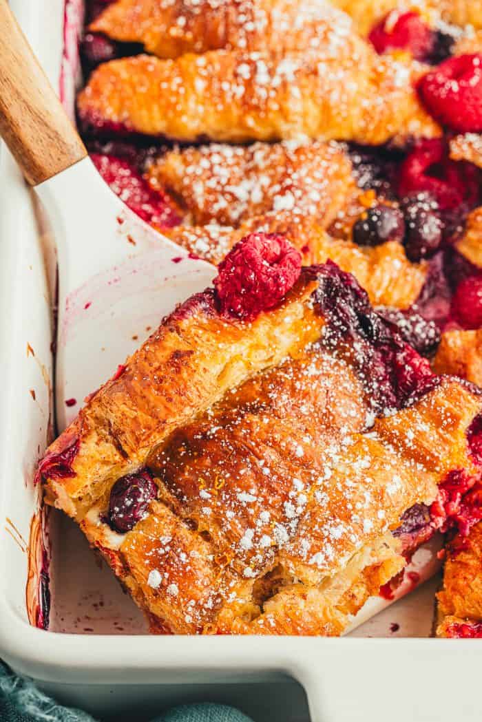 Croissant French toast bake in a white casserole dish with a spatula removing a slice of it.