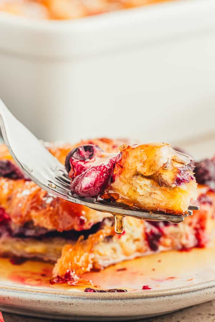 A white plate with a slice of croissant French toast bake that is covered in maple syrup and a fork with a bite of the casserole.