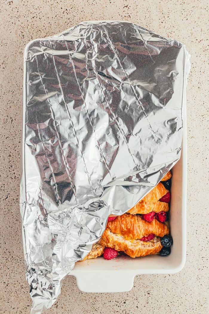 A white baking tray with sliced croissants and raspberries and blueberries and aluminum foil.