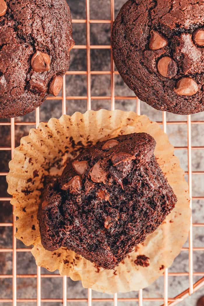 A close up of a double chocolate muffin with a bite take out of it, laying on a cooling rack with two other muffins.