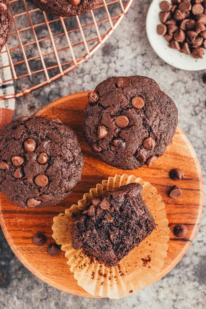 A wooden board with three double chocolate muffins and one is on its side with a bite taken out of it.