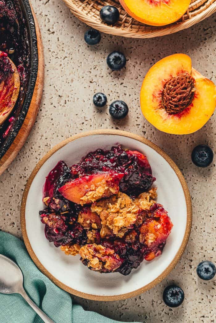 A bowl of peach crumble with a half of a ripe peach and blueberries scattered around. 