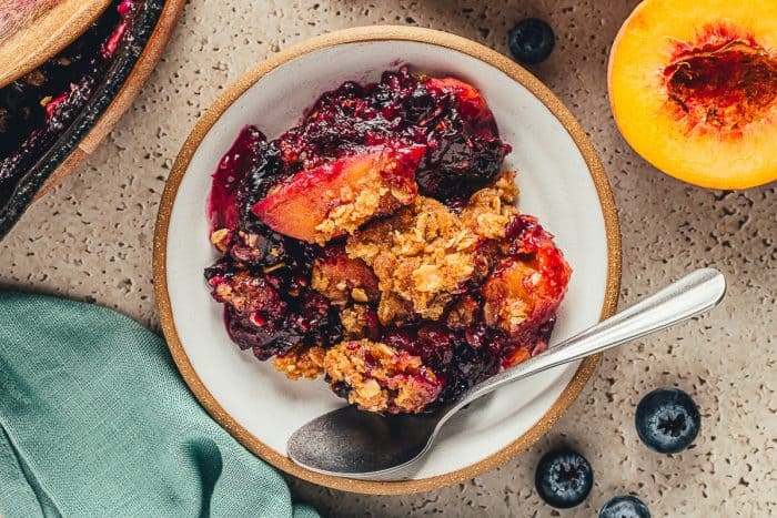A bowl of blueberry peach crumble with a spoon and a peach cut in half.