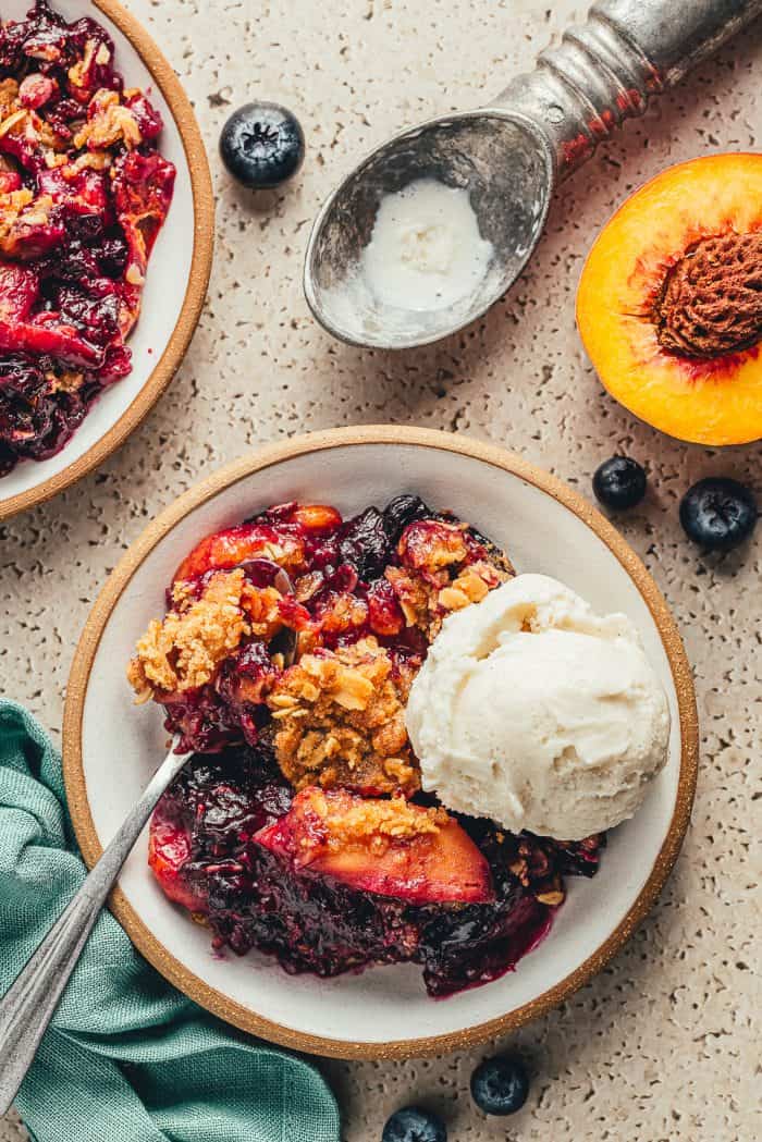 A white bowl with blueberry peach crumble and a scoop of vanilla ice cream.