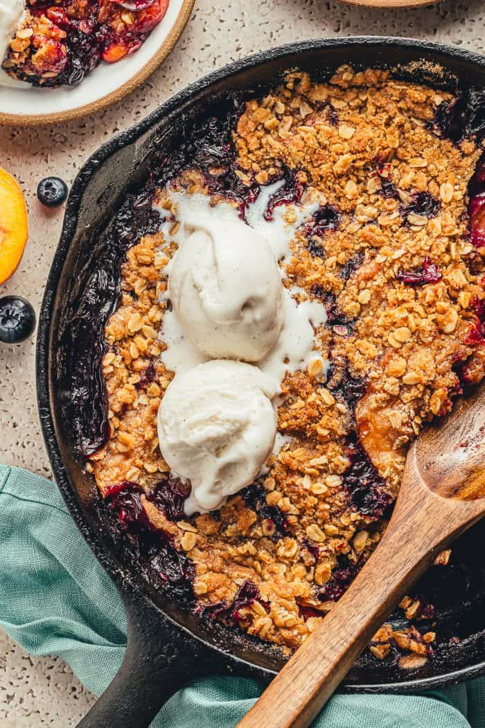 A skillet with a blueberry peach crumble garnished with two scoops of ice cream and a wooden spoon.