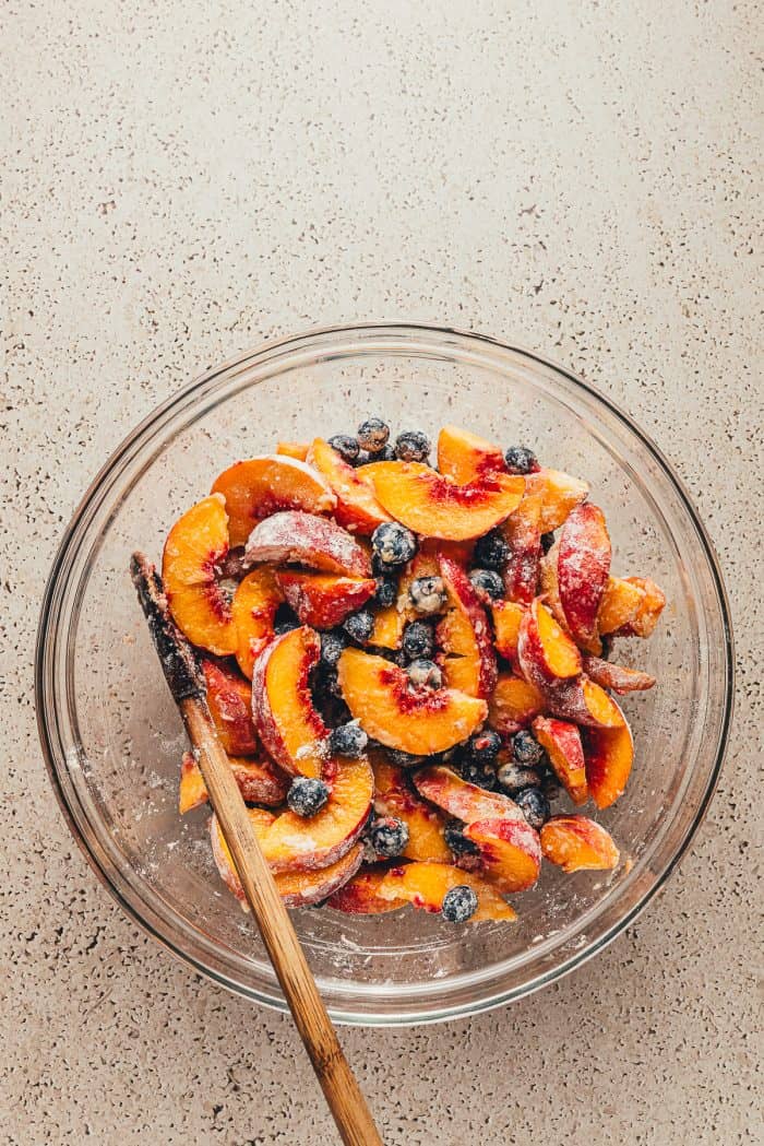 A glass bowl with peaches and blueberries that have been tossed in sugar and flour.